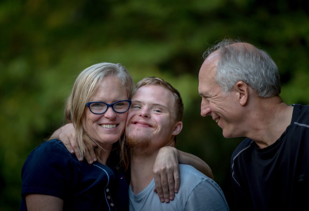 Das Bild zeigt eine Familie (Mutter und Vater) die ihren Sohn mit Downsyndrom umarmen.